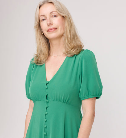 A woman with blonde hair wears the Robyn Green Button Through Midi Dress, which features an empire waist, puff sleeves, and fabric-covered buttons. She stands against a plain light background, smiling softly at the camera.