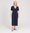 A woman with long, light blonde hair smiles while standing in the Robyn Navy Button Through Midi Dress with short sleeves and an empire waist, paired with blue low-heeled shoes against a plain light background.
