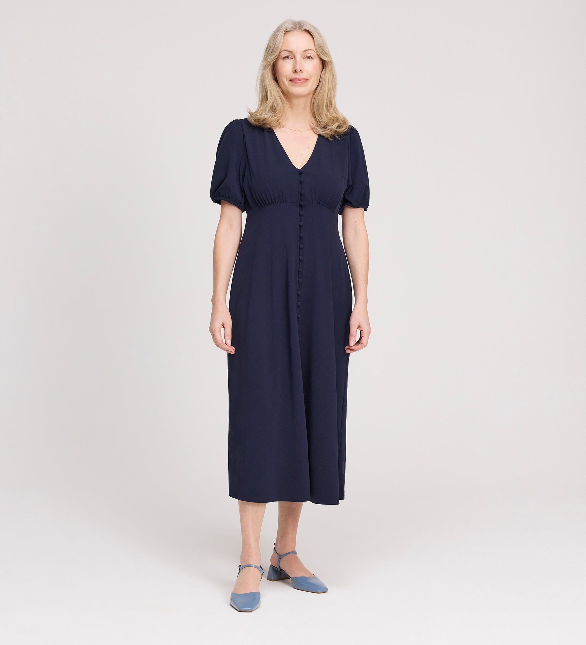 A woman with long, light blonde hair smiles while standing in the Robyn Navy Button Through Midi Dress with short sleeves and an empire waist, paired with blue low-heeled shoes against a plain light background.