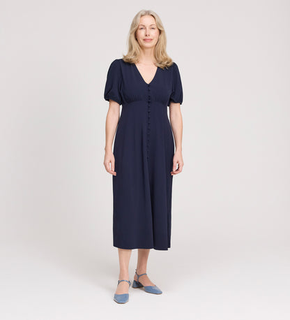 A woman with long, light blonde hair smiles while standing in the Robyn Navy Button Through Midi Dress with short sleeves and an empire waist, paired with blue low-heeled shoes against a plain light background.
