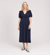 A woman with long, light hair stands in a studio, wearing the Robyn Navy Button Through Midi Dress with short sleeves and an empire waist. She faces forward, smiles slightly, and pairs the dress with blue strap shoes against a neutral background.