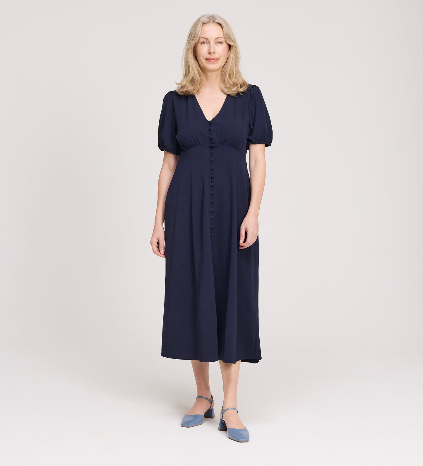 A woman with long, light hair stands in a studio, wearing the Robyn Navy Button Through Midi Dress with short sleeves and an empire waist. She faces forward, smiles slightly, and pairs the dress with blue strap shoes against a neutral background.