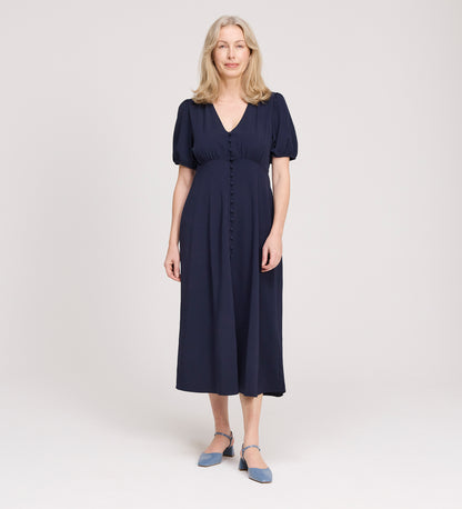 A woman with long, light hair stands in a studio, wearing the Robyn Navy Button Through Midi Dress with short sleeves and an empire waist. She faces forward, smiles slightly, and pairs the dress with blue strap shoes against a neutral background.