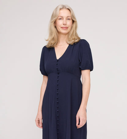 A woman with shoulder-length blonde hair wears the Robyn Navy Button Through Midi Dress with short puff sleeves and an empire waist, standing against a plain light background and smiling softly at the camera.
