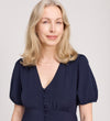 A woman with shoulder-length blonde hair, in the Robyn Navy Button Through Midi Dress, featuring puffed short sleeves, an empire waist, and a V-neckline, smiles softly against a light neutral background.