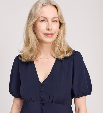 A woman with shoulder-length blonde hair, in the Robyn Navy Button Through Midi Dress, featuring puffed short sleeves, an empire waist, and a V-neckline, smiles softly against a light neutral background.