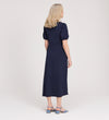 A woman with shoulder-length blonde hair stands facing away, wearing the Robyn Navy Button Through Midi Dress and blue heeled sandals against a plain light background.