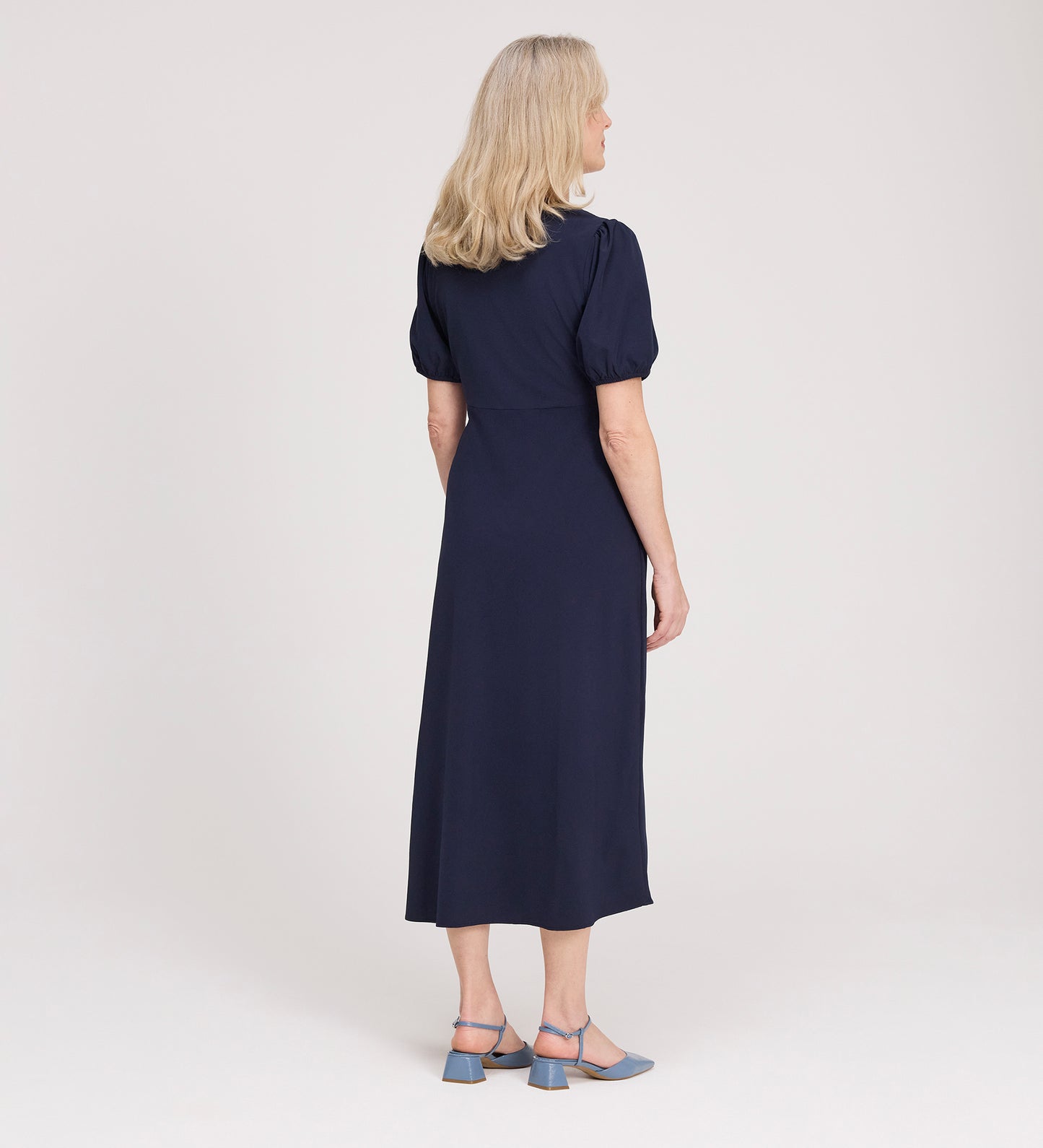 A woman with shoulder-length blonde hair stands facing away, wearing the Robyn Navy Button Through Midi Dress and blue heeled sandals against a plain light background.