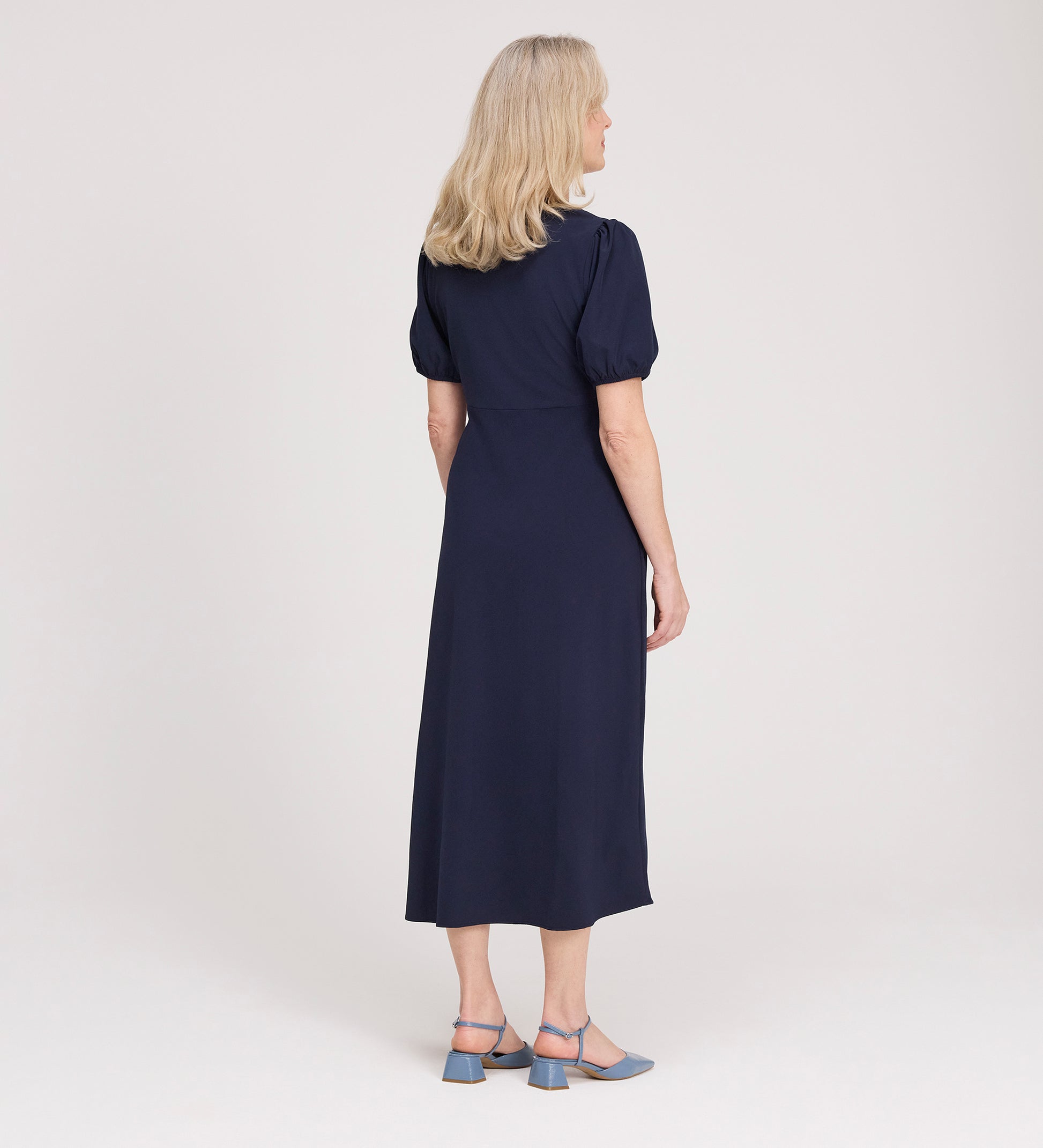 A woman with shoulder-length blonde hair stands facing away, wearing the Robyn Navy Button Through Midi Dress and blue heeled sandals against a plain light background.