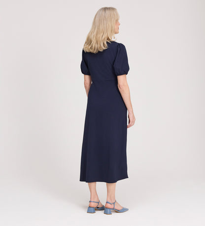 A woman with shoulder-length blonde hair stands facing away, wearing the Robyn Navy Button Through Midi Dress and blue heeled sandals against a plain light background.