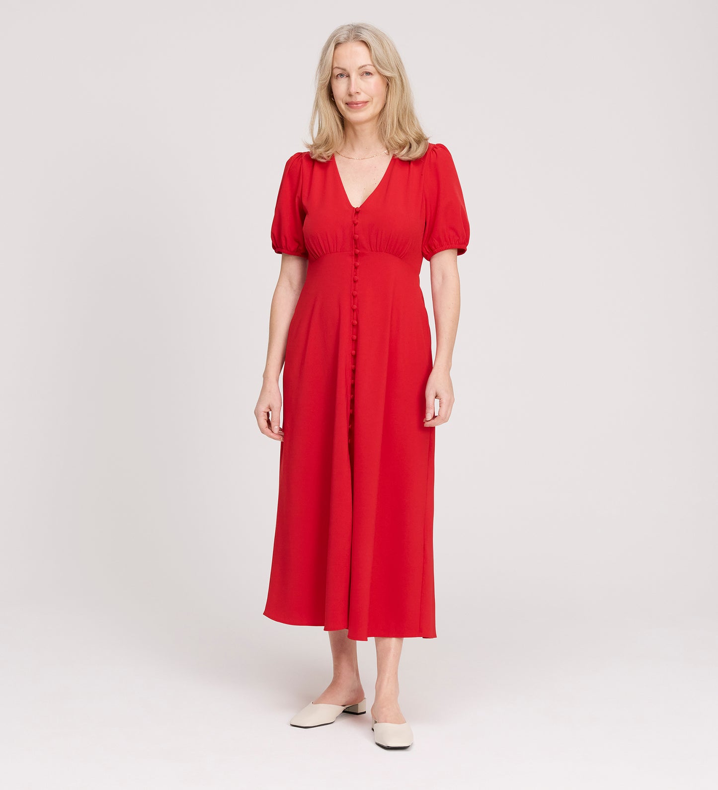 A woman with light blonde hair smiles softly as she stands against a plain white background, wearing the Robyn Red Button Through Midi Dress with short sleeves and white flats.