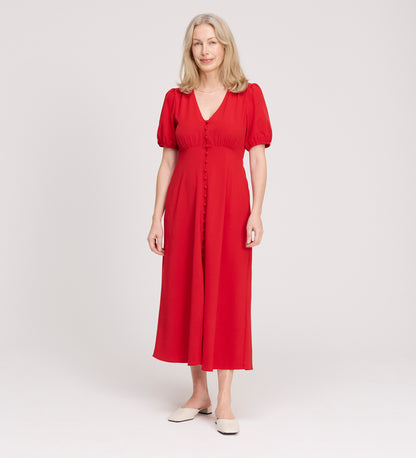 A woman with light blonde hair smiles softly as she stands against a plain white background, wearing the Robyn Red Button Through Midi Dress with short sleeves and white flats.