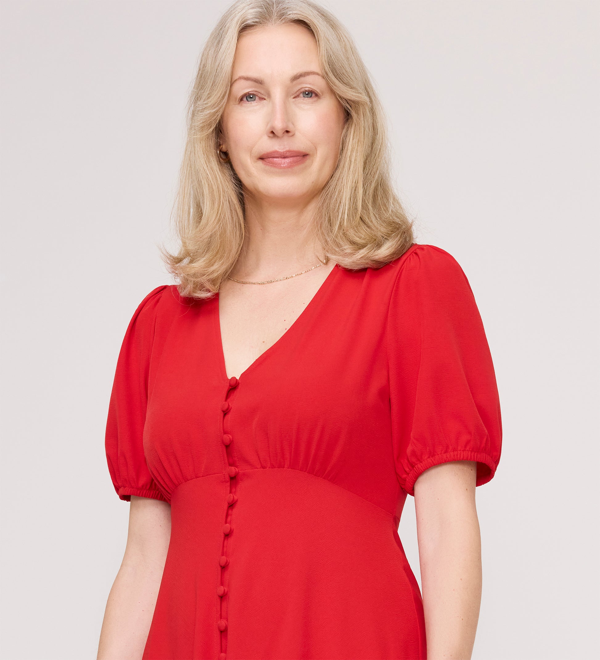 A blonde woman wears the Robyn Red Button Through Midi Dress with puff sleeves and fabric-covered buttons, standing against a plain light background and smiling gently—a perfect day-to-evening look.