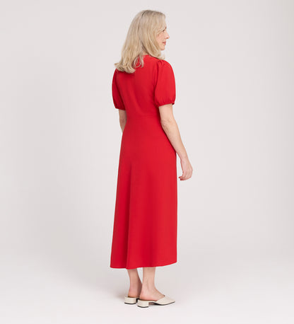 A woman with blonde hair stands facing away in a light studio, wearing the Robyn Red Button Through Midi Dress with short sleeves and beige slip-on shoes—an effortless look for day to evening.