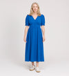 A woman with long, light blonde hair is wearing the Samantha Cobalt Button Through Midi Dress and white sneakers, standing against a plain light background, facing forward with a slight smile.