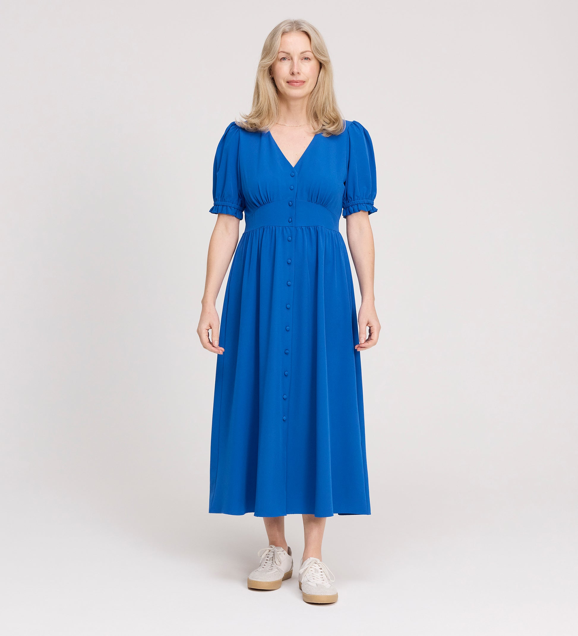 A woman with long, light blonde hair is wearing the Samantha Cobalt Button Through Midi Dress and white sneakers, standing against a plain light background, facing forward with a slight smile.