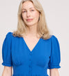 A woman with long blonde hair models the Samantha Cobalt Button Through Midi Dress, featuring short sleeves and front buttons, as she poses gently against a plain light background.