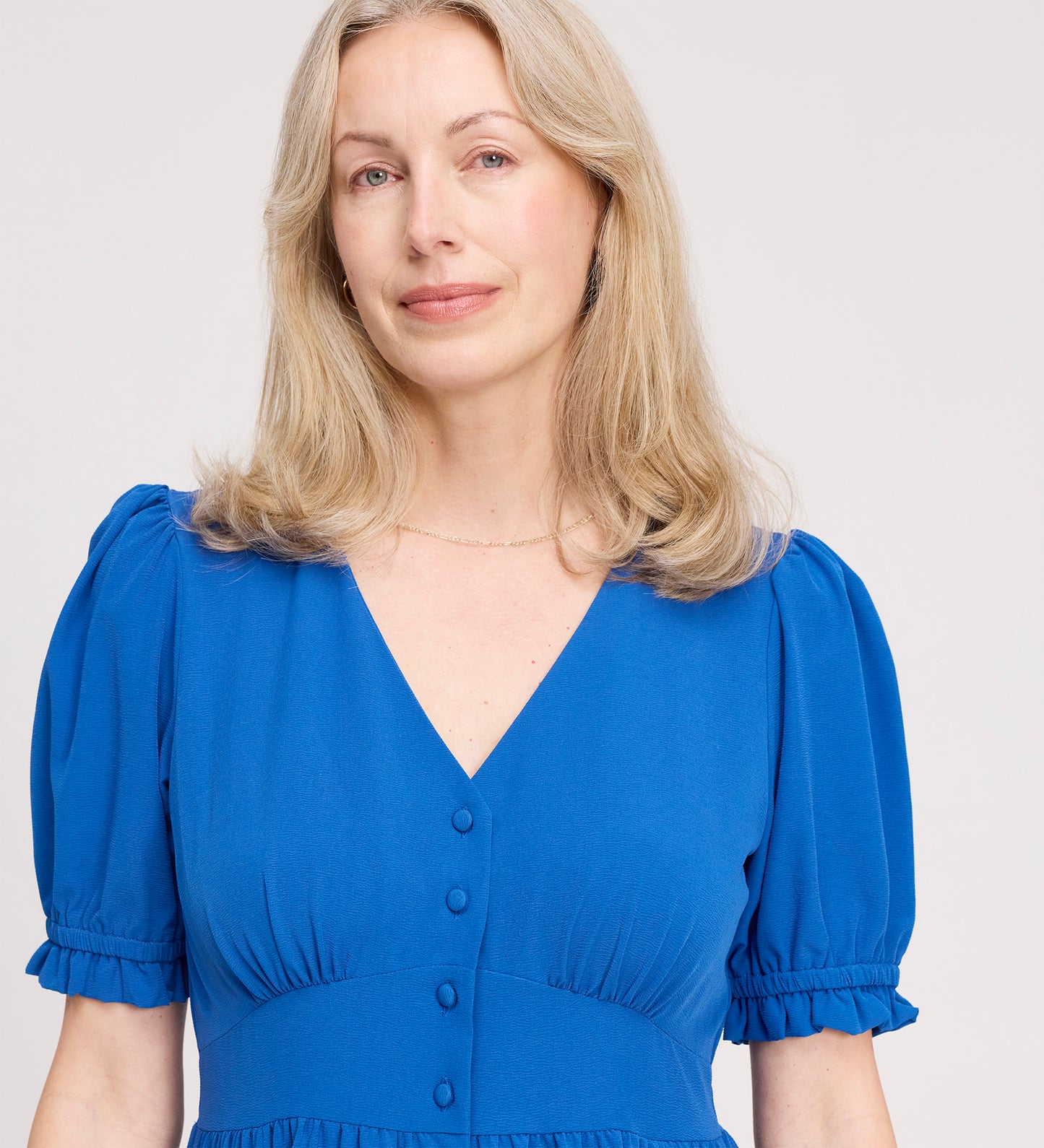 A woman with long blonde hair models the Samantha Cobalt Button Through Midi Dress, featuring short sleeves and front buttons, as she poses gently against a plain light background.