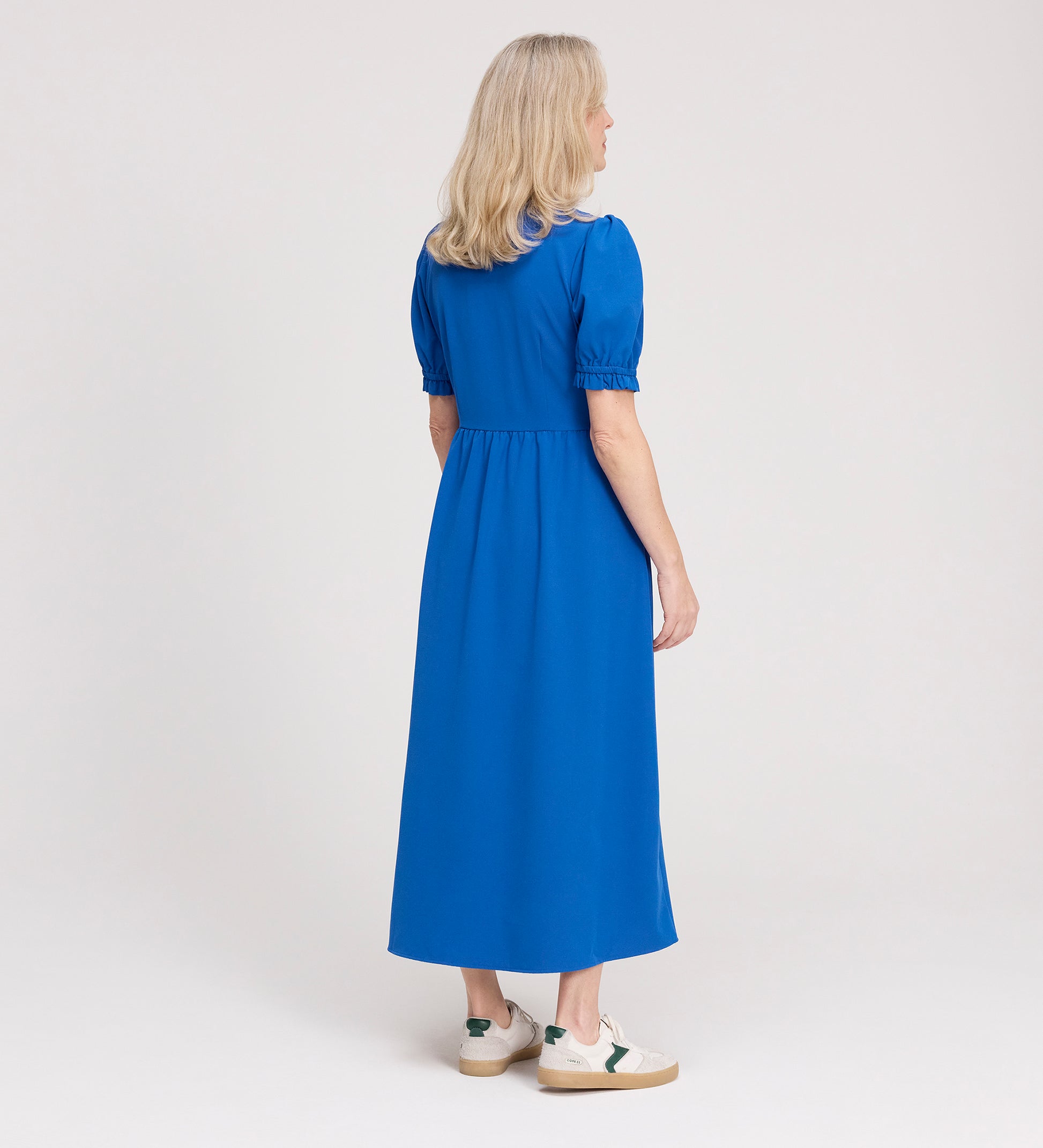 A woman with shoulder-length blonde hair faces away, wearing the Samantha Cobalt Button Through Midi Dress and white sneakers with green accents, set against a plain light background.