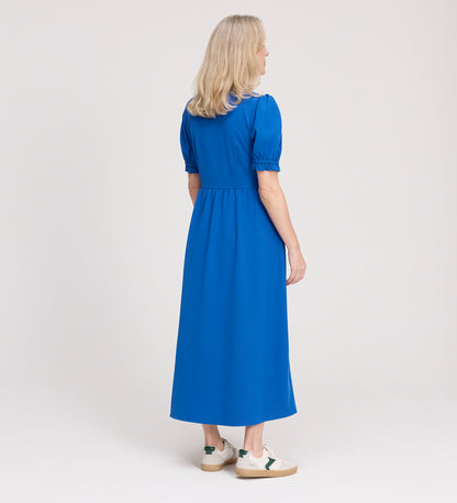 A woman with shoulder-length blonde hair faces away, wearing the Samantha Cobalt Button Through Midi Dress and white sneakers with green accents, set against a plain light background.