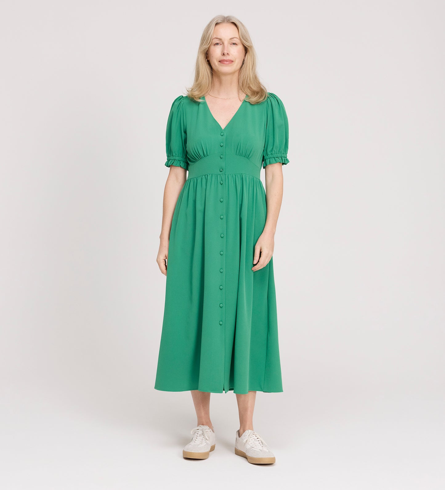 A woman with long, light blond hair stands against a plain background, wearing the Samantha Green Button Through Midi Dress with puff sleeves and white sneakers. She looks straight ahead with a neutral expression.