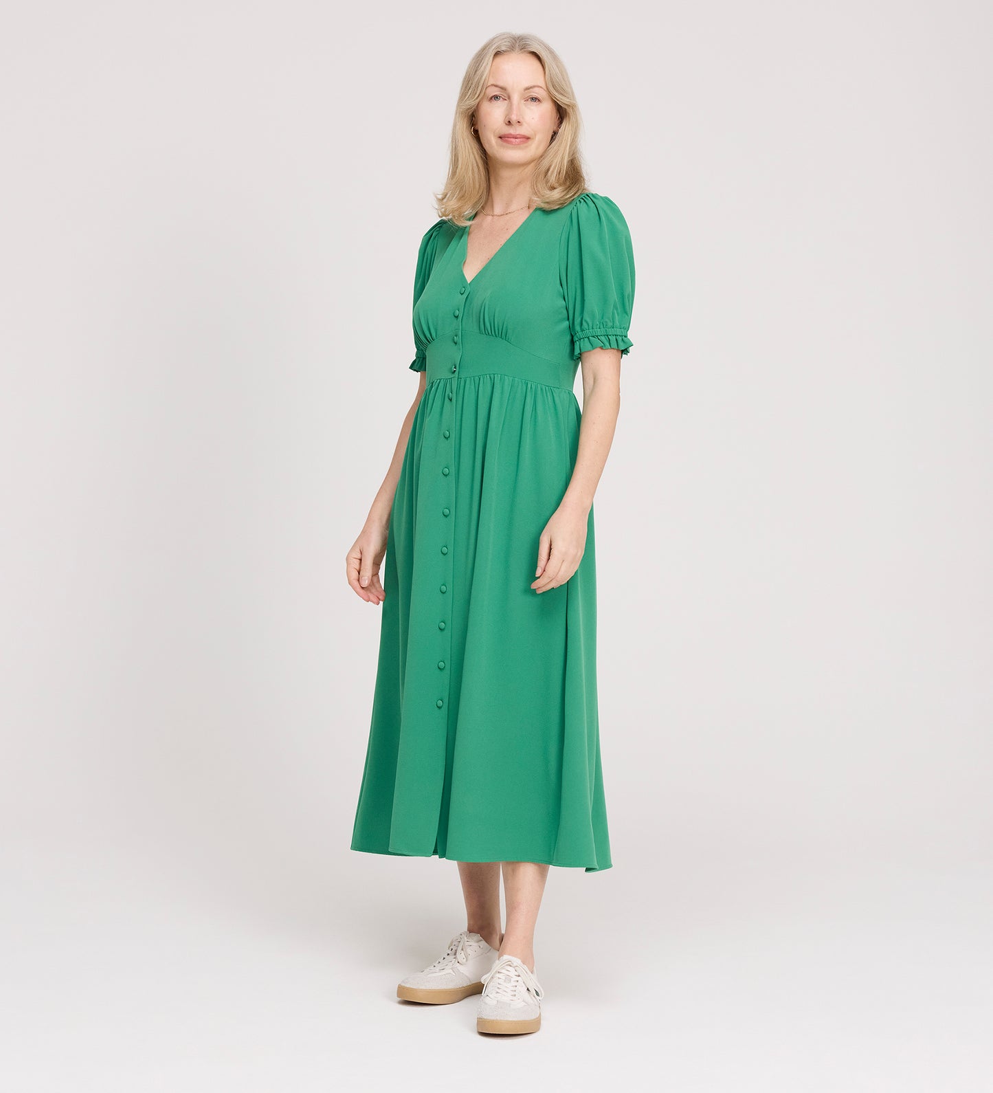 A woman with light hair stands against a plain background, wearing the Samantha Green Button Through Midi Dress with white sneakers. She looks at the camera and smiles slightly.
