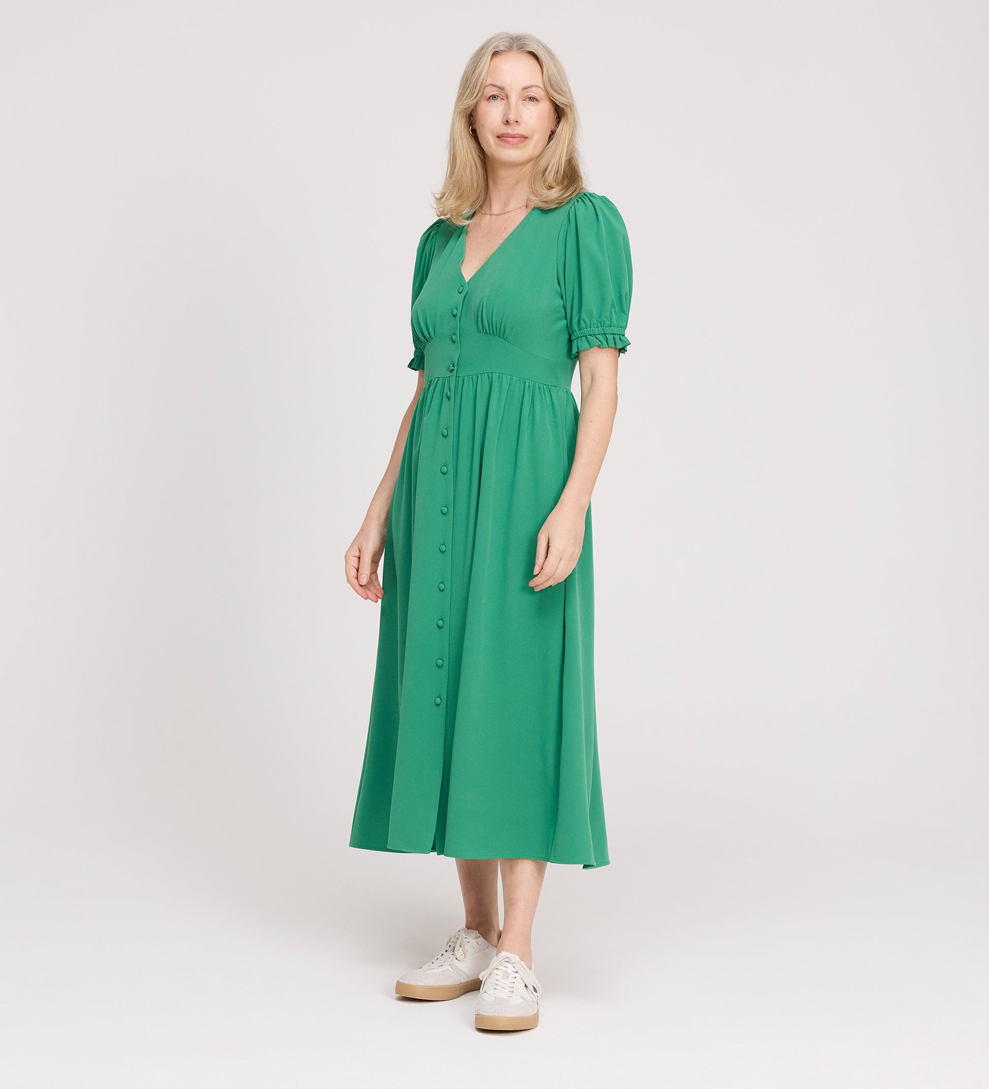 A woman with light hair stands against a plain background, wearing the Samantha Green Button Through Midi Dress with white sneakers. She looks at the camera and smiles slightly.