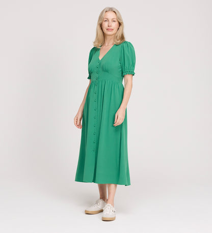 A woman with light hair stands against a plain background, wearing the Samantha Green Button Through Midi Dress with white sneakers. She looks at the camera and smiles slightly.
