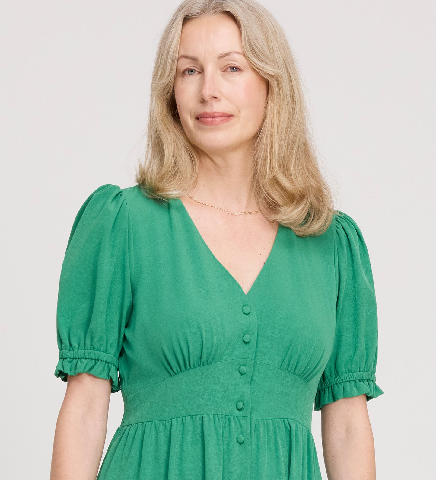 A woman with wavy blonde hair, smiling gently, wears the Samantha Green Button Through Midi Dress with a V-neckline and short puff sleeves, standing against a plain light background.