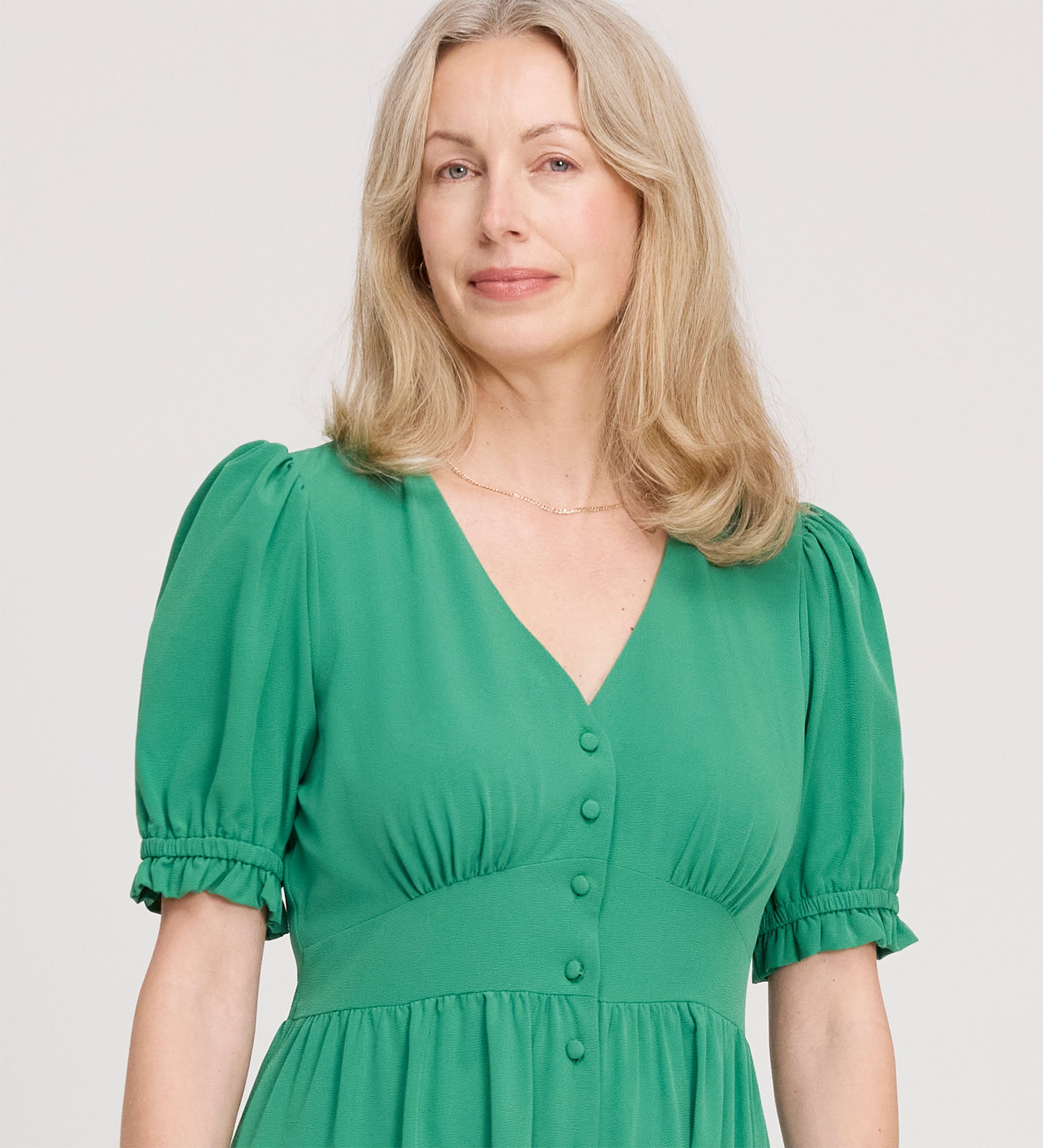 A woman with wavy blonde hair, smiling gently, wears the Samantha Green Button Through Midi Dress with a V-neckline and short puff sleeves, standing against a plain light background.