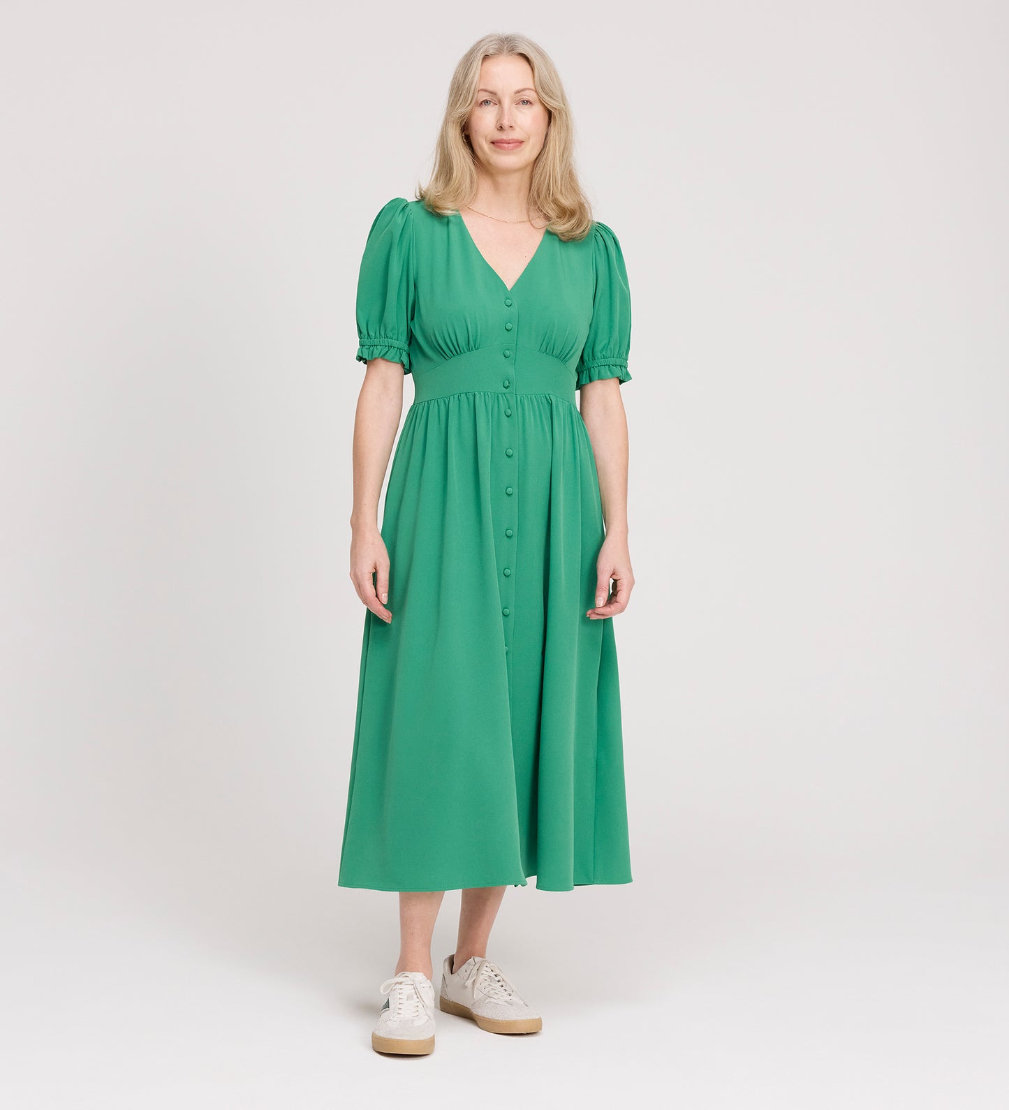 A woman with long, light hair wears the Samantha Green Button Through Midi Dress, featuring puff sleeves and a V-neckline, paired with white sneakers, standing against a plain white background.
