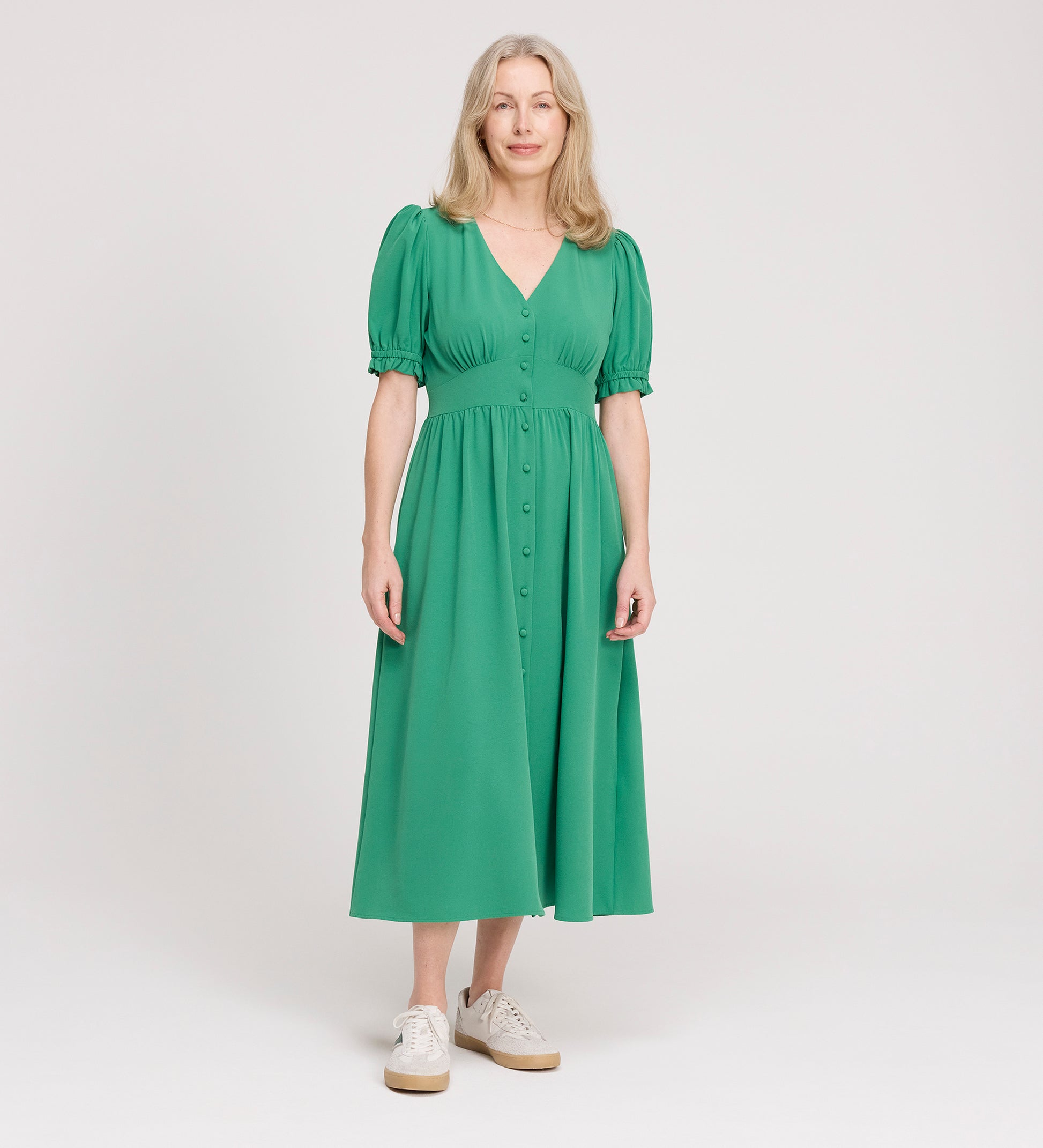 A woman with long, light hair wears the Samantha Green Button Through Midi Dress, featuring puff sleeves and a V-neckline, paired with white sneakers, standing against a plain white background.