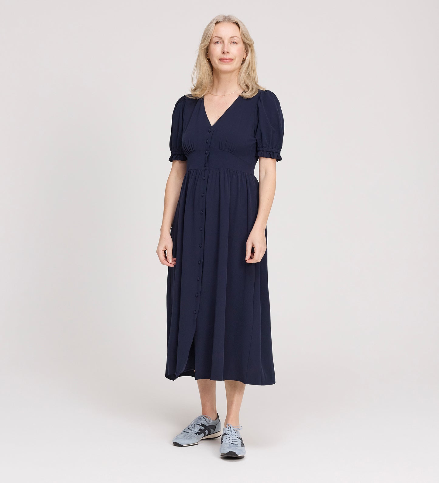 A woman with light hair stands in a studio, smiling slightly and facing forward, wearing the Samantha Navy Button Through Midi Dress with puff sleeves and a V-neck, paired with gray sneakers against a plain light background.