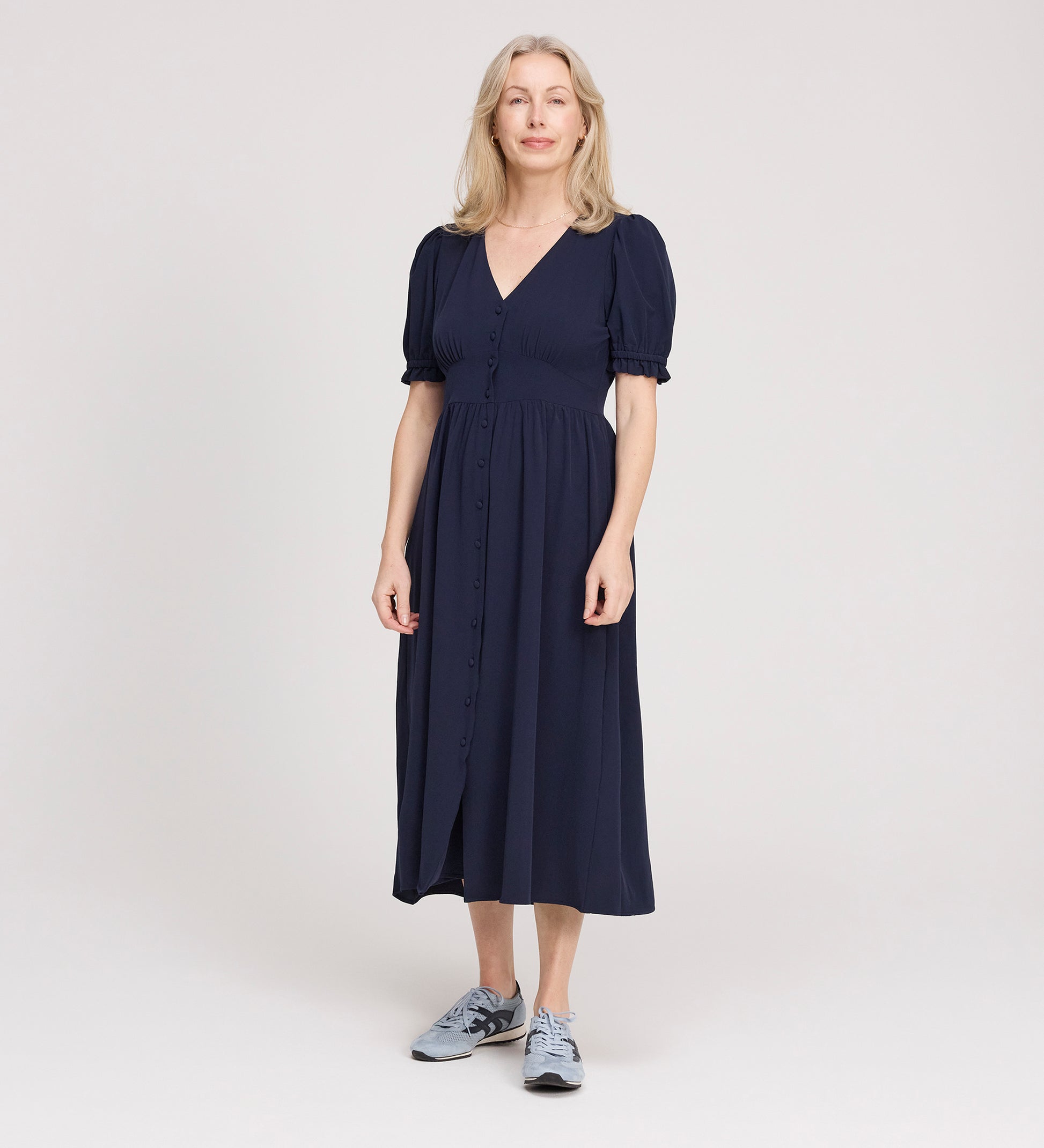 A woman with light hair stands in a studio, smiling slightly and facing forward, wearing the Samantha Navy Button Through Midi Dress with puff sleeves and a V-neck, paired with gray sneakers against a plain light background.