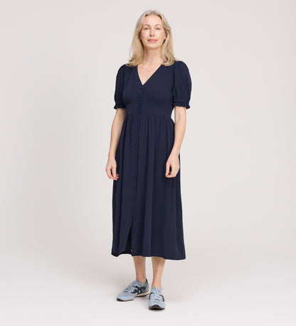 A woman with light hair stands in a studio, smiling slightly and facing forward, wearing the Samantha Navy Button Through Midi Dress with puff sleeves and a V-neck, paired with gray sneakers against a plain light background.