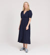 A woman with light hair wears the Samantha Navy Button Through Midi Dress, featuring short puff sleeves and a V-neck, paired with grey sneakers, standing against a plain light background.