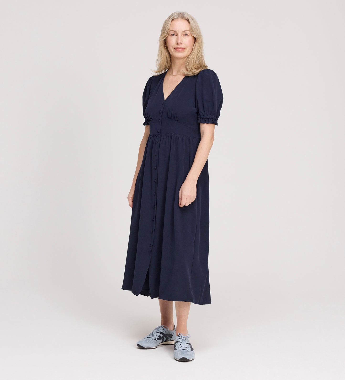 A woman with light hair wears the Samantha Navy Button Through Midi Dress, featuring short puff sleeves and a V-neck, paired with grey sneakers, standing against a plain light background.