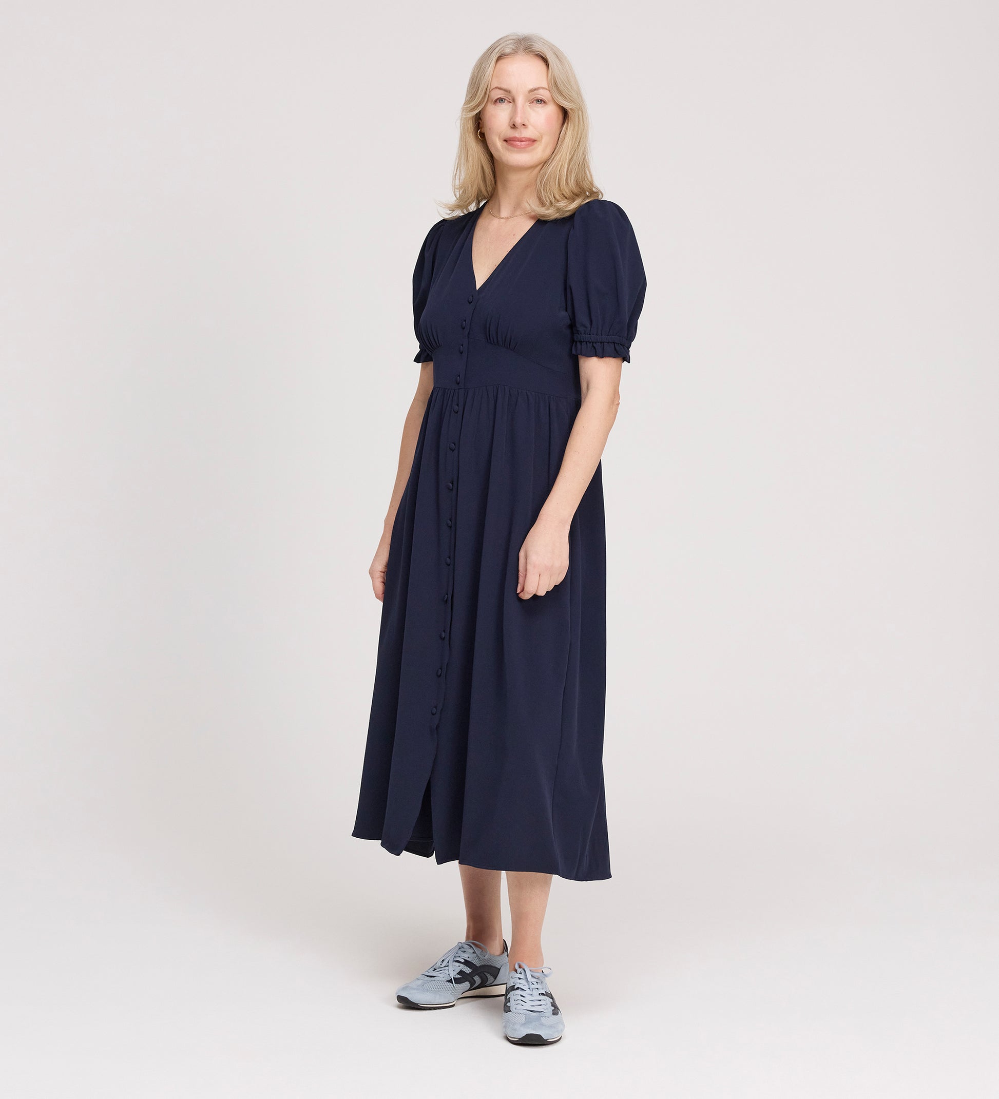 A woman with light hair wears the Samantha Navy Button Through Midi Dress, featuring short puff sleeves and a V-neck, paired with grey sneakers, standing against a plain light background.