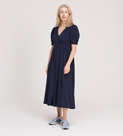 A woman with light hair wears the Samantha Navy Button Through Midi Dress, featuring short puff sleeves and a V-neck, paired with grey sneakers, standing against a plain light background.