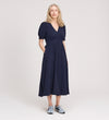 A woman with light blonde hair smiles slightly, standing against a plain light background with her hands in her pockets, wearing the Samantha Navy Button Through Midi Dress and light blue sneakers.