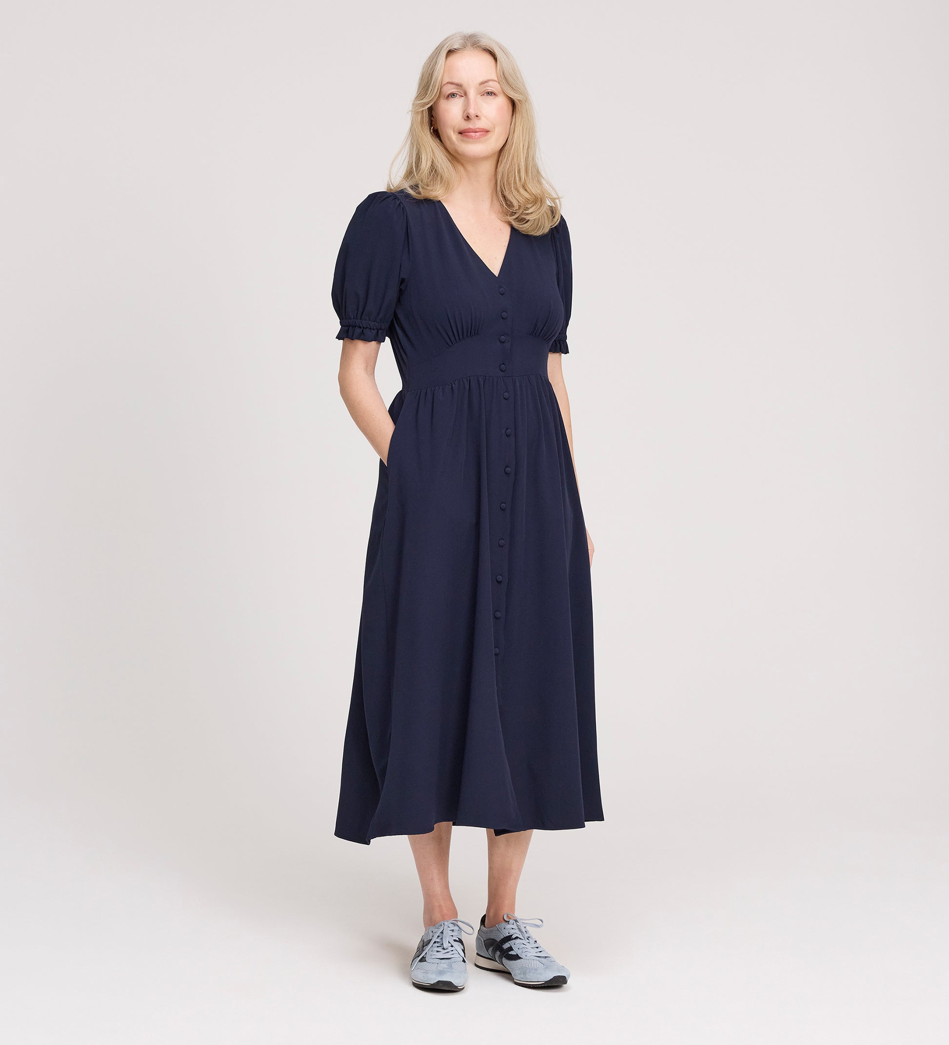 A woman with light blonde hair smiles slightly, standing against a plain light background with her hands in her pockets, wearing the Samantha Navy Button Through Midi Dress and light blue sneakers.