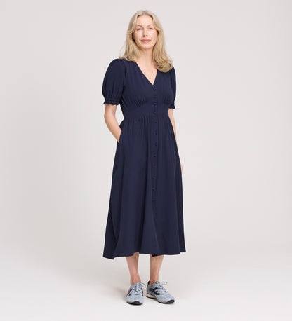 A woman with light blonde hair smiles slightly, standing against a plain light background with her hands in her pockets, wearing the Samantha Navy Button Through Midi Dress and light blue sneakers.