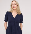 A woman with light blonde hair smiles softly, standing against a plain light background with her hands in her pockets while wearing the Samantha Navy Button Through Midi Dress.