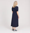 A woman with blonde hair faces away, wearing the Samantha Navy Button Through Midi Dress and gray sneakers with navy accents, set against a plain light background.
