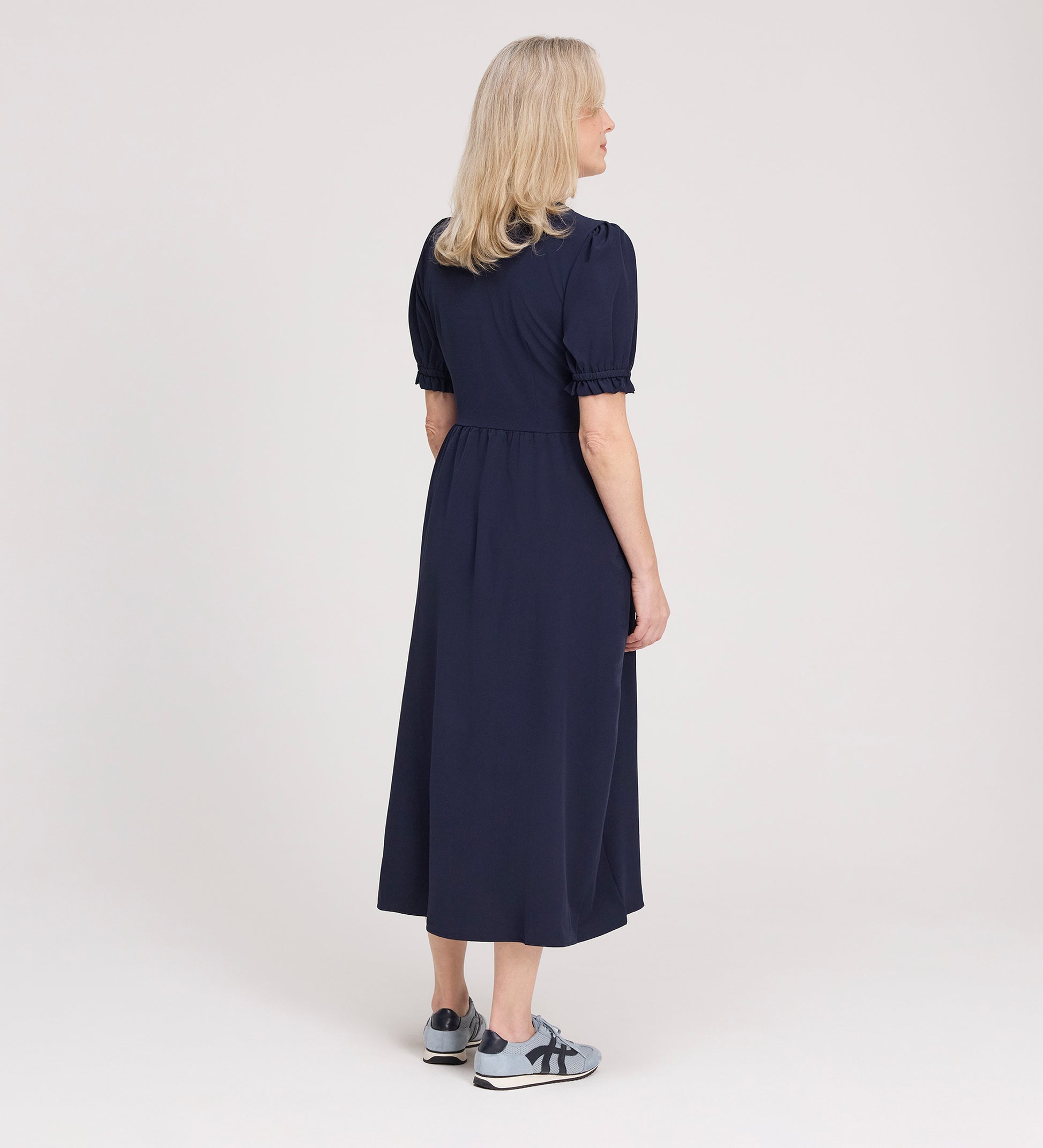 A woman with blonde hair faces away, wearing the Samantha Navy Button Through Midi Dress and gray sneakers with navy accents, set against a plain light background.