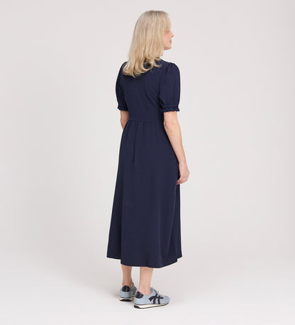 A woman with blonde hair faces away, wearing the Samantha Navy Button Through Midi Dress and gray sneakers with navy accents, set against a plain light background.