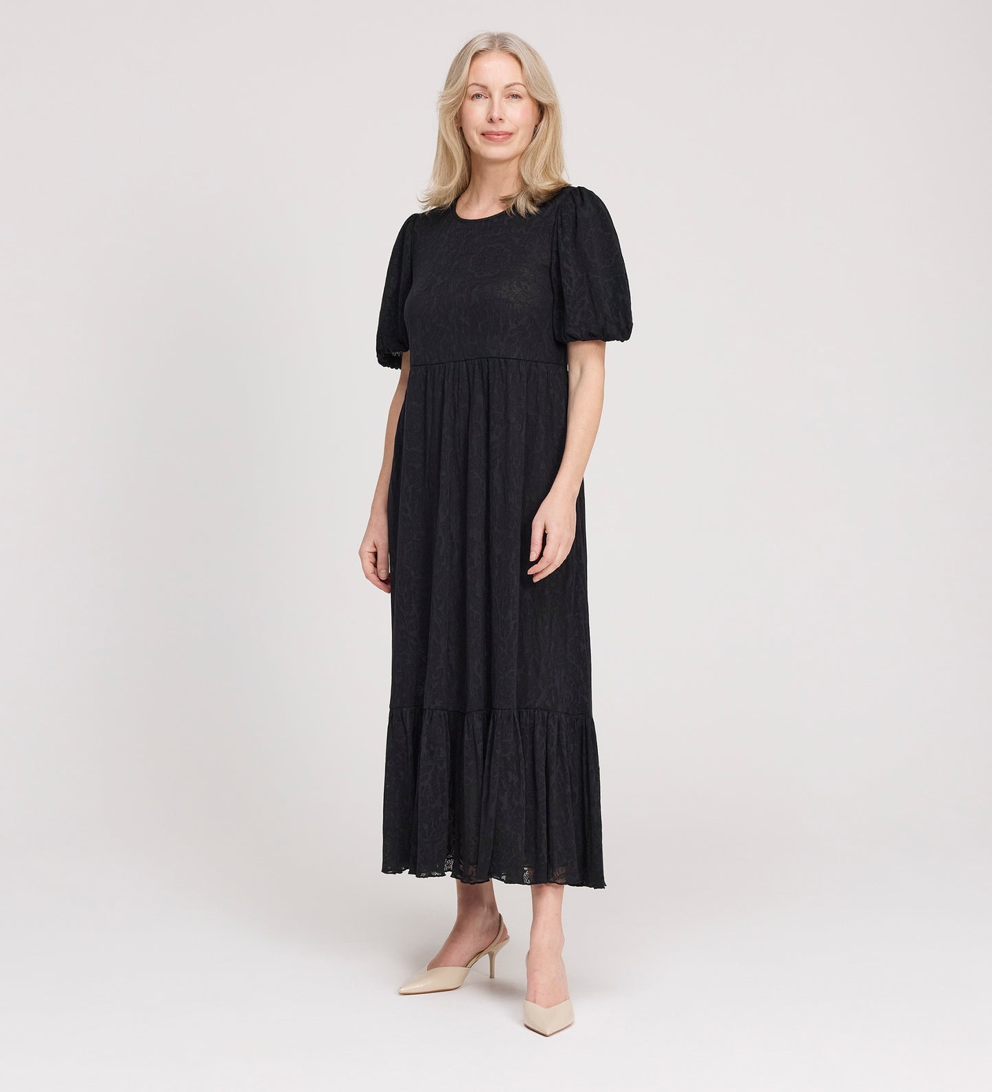 A woman with long blonde hair is standing and smiling, wearing the Yasmine Black Jersey Midi Dress with puff sleeves and a flared hem, paired with light-colored heels, against a plain white background.