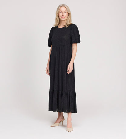 A woman with long blonde hair is standing and smiling, wearing the Yasmine Black Jersey Midi Dress with puff sleeves and a flared hem, paired with light-colored heels, against a plain white background.