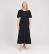 A woman with light hair stands against a plain background, wearing the Yasmine Black Jersey Midi Dress with puff sleeves and a ruffled hem, paired with beige pointed-toe heels.