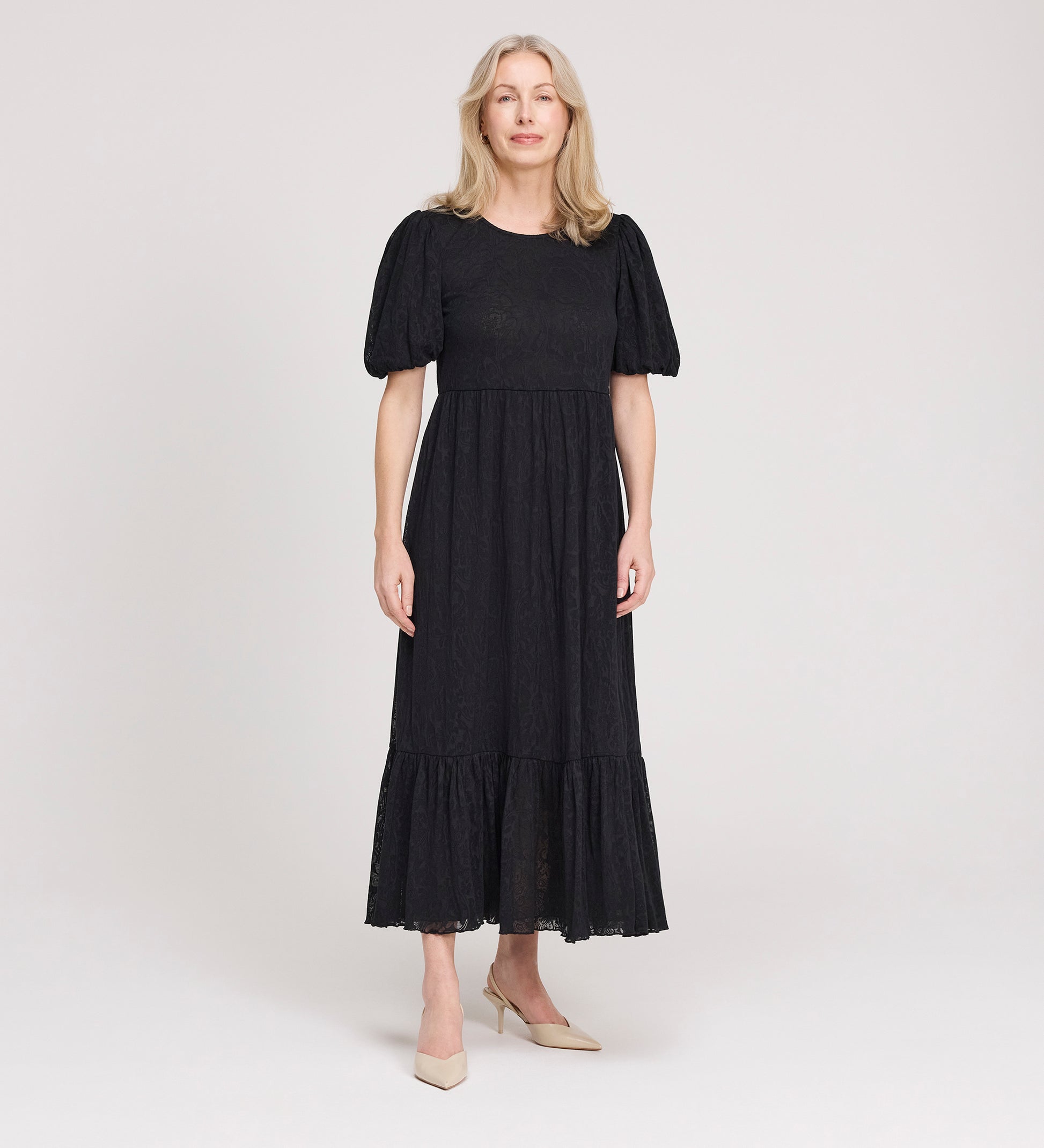 A woman with light hair stands against a plain background, wearing the Yasmine Black Jersey Midi Dress with puff sleeves and a ruffled hem, paired with beige pointed-toe heels.
